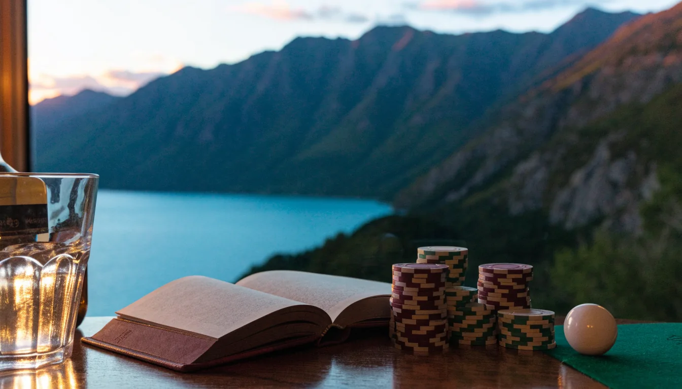 Open guidebook and casino chips on a polished table beside a window overlooking New Zealand Alps at sunset.